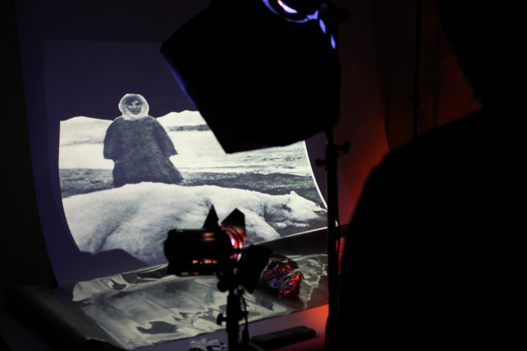 an installation image of a filming set, we can see a camera and lighting set up, behind this there is a projection of a person of Inupiat origin