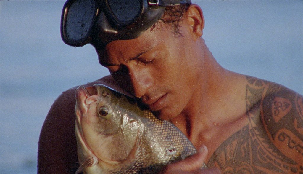 A man with a snorkle on his head holds a fish against his bare chest in a loving embrace